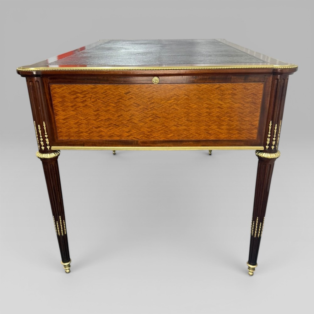 A Louis XVI style mahogany and parquetry writing desk featuring a dark leather top, gilt-bronze mounts, five drawers, and fluted tapered legs. View IX