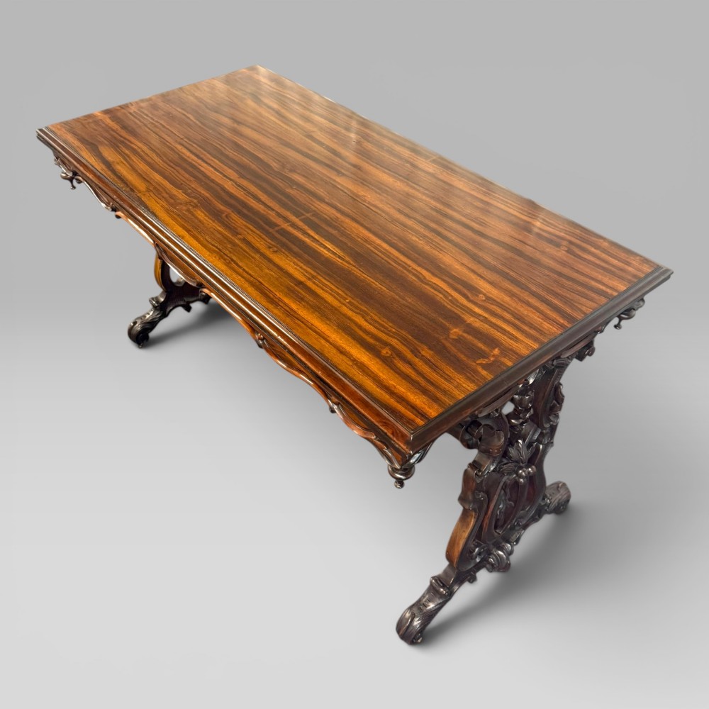 An Early Victorian rectangular centre table featuring a figured zebrawood veneered top, carved aprons, and carved rosewood trestle standards with scrolled feet.