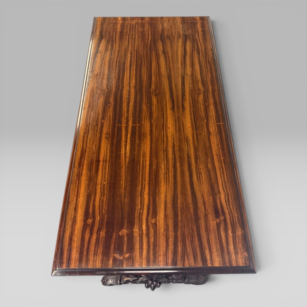 An Early Victorian rectangular centre table featuring a figured zebrawood veneered top, carved aprons, and carved rosewood trestle standards with scrolled feet. View V