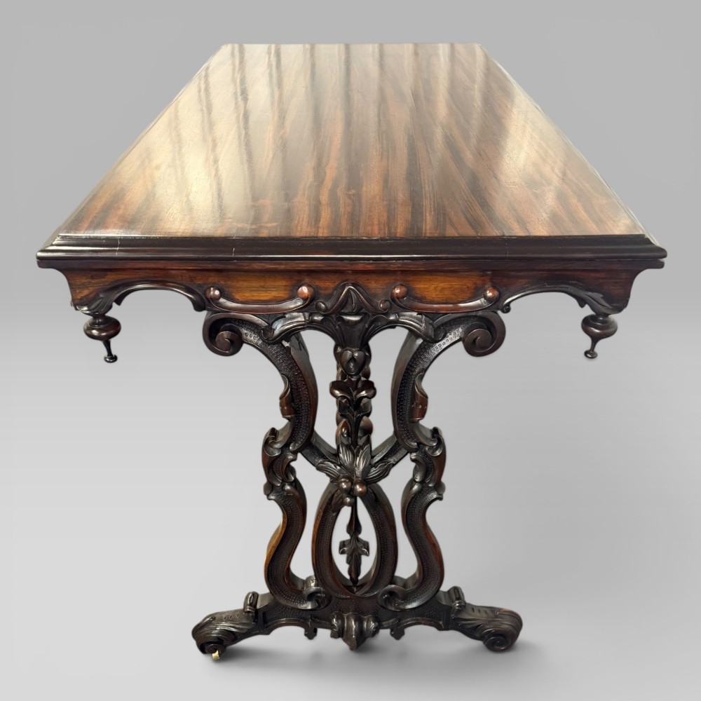 An Early Victorian rectangular centre table featuring a figured zebrawood veneered top, carved aprons, and carved rosewood trestle standards with scrolled feet. View VI