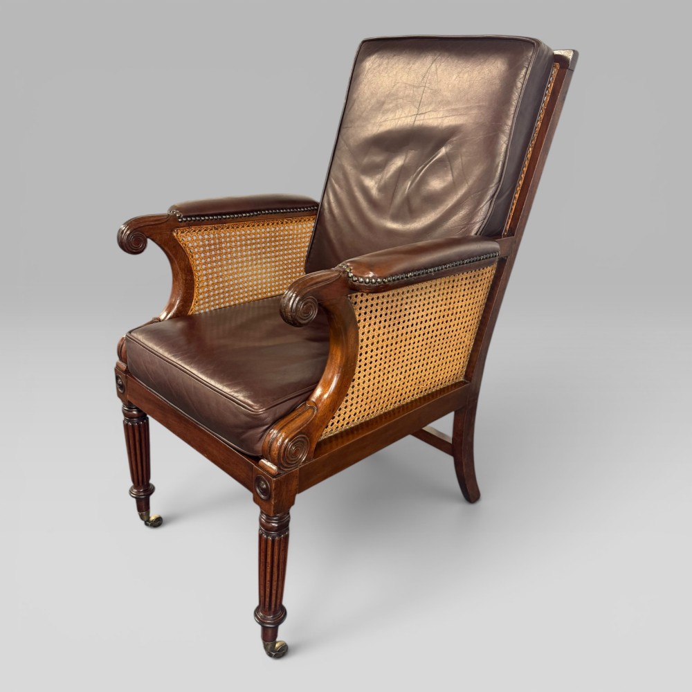 A Regency mahogany bergere armchair featuring dark brown leather cushions, woven cane side panels, scroll arms, and reeded front legs with brass castors.