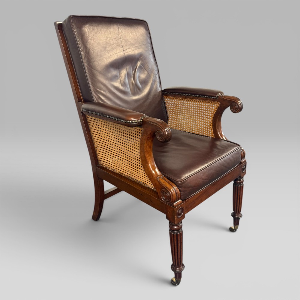 A Regency mahogany bergere armchair featuring dark brown leather cushions, woven cane side panels, scroll arms, and reeded front legs with brass castors. View II