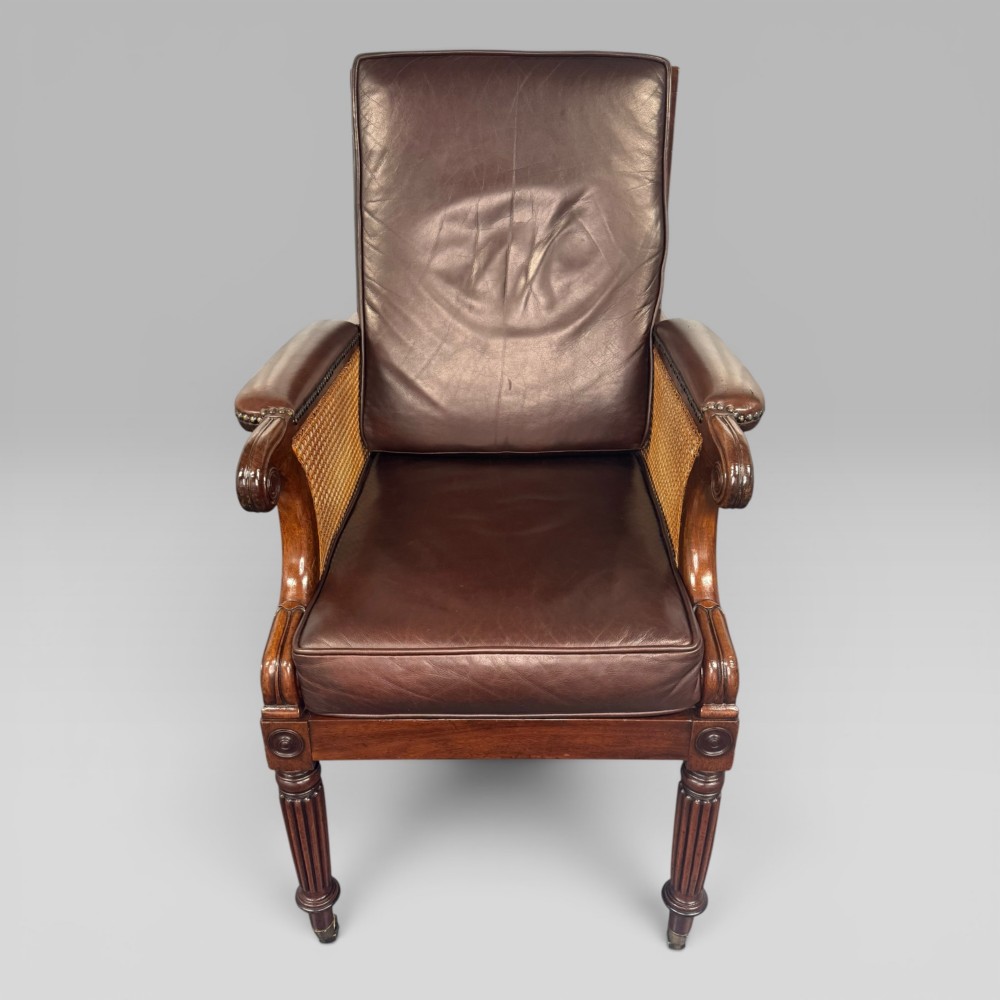 A Regency mahogany bergere armchair featuring dark brown leather cushions, woven cane side panels, scroll arms, and reeded front legs with brass castors. View III