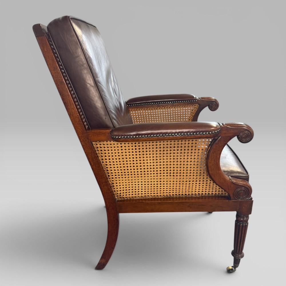A Regency mahogany bergere armchair featuring dark brown leather cushions, woven cane side panels, scroll arms, and reeded front legs with brass castors. View IV