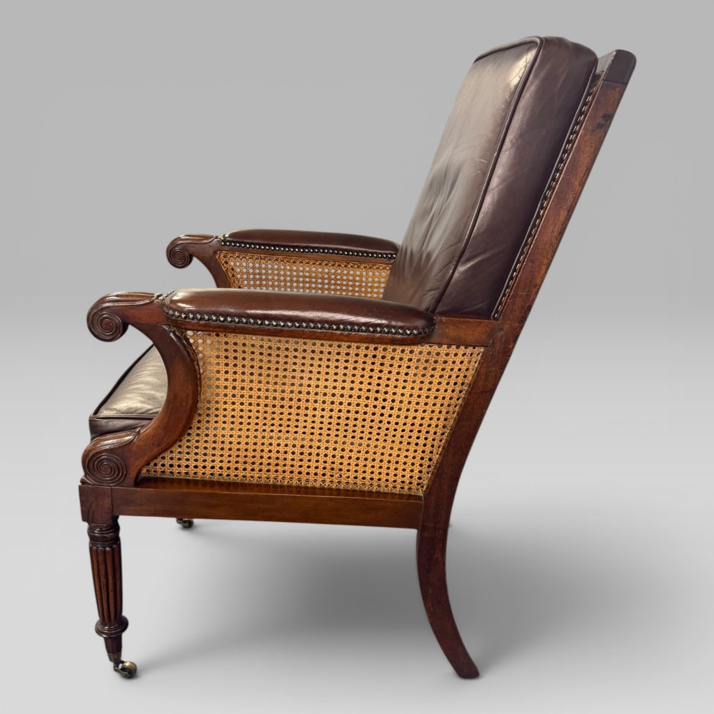 A Regency mahogany bergere armchair featuring dark brown leather cushions, woven cane side panels, scroll arms, and reeded front legs with brass castors. View V