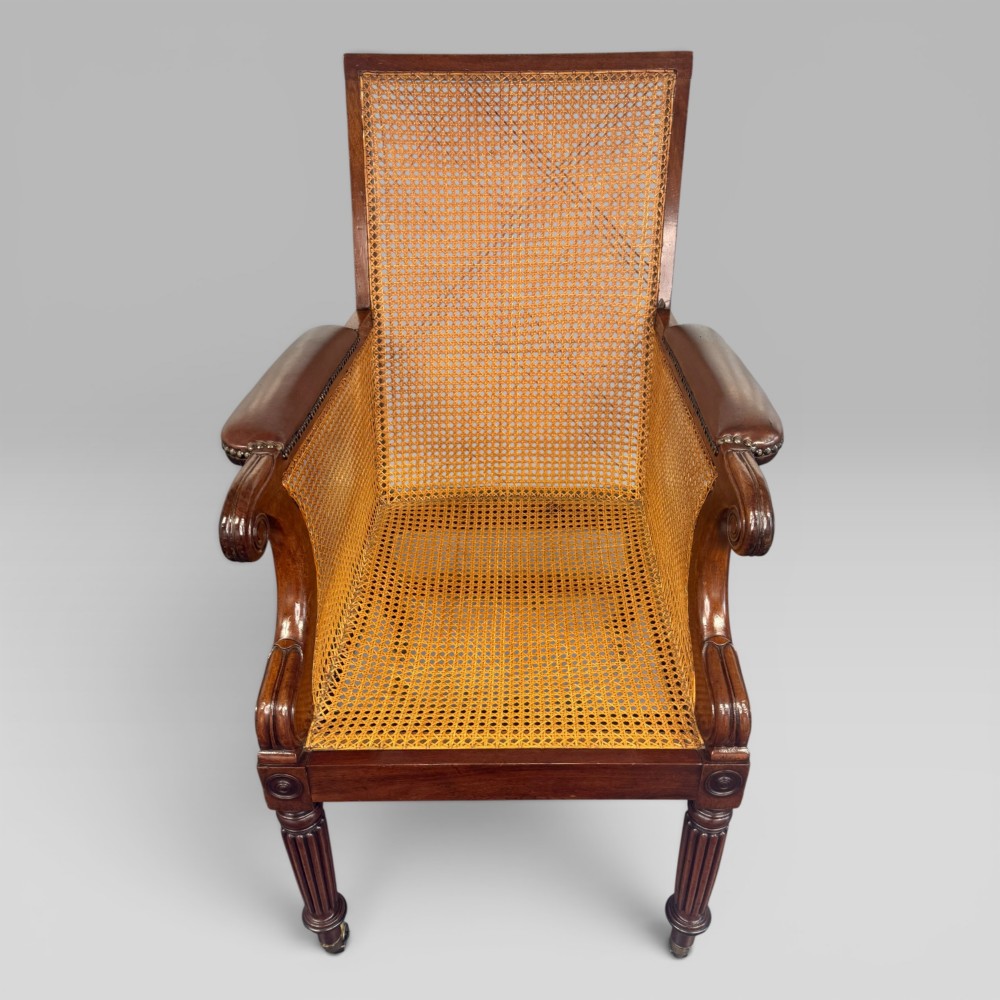 A Regency mahogany bergere armchair featuring dark brown leather cushions, woven cane side panels, scroll arms, and reeded front legs with brass castors. View VI