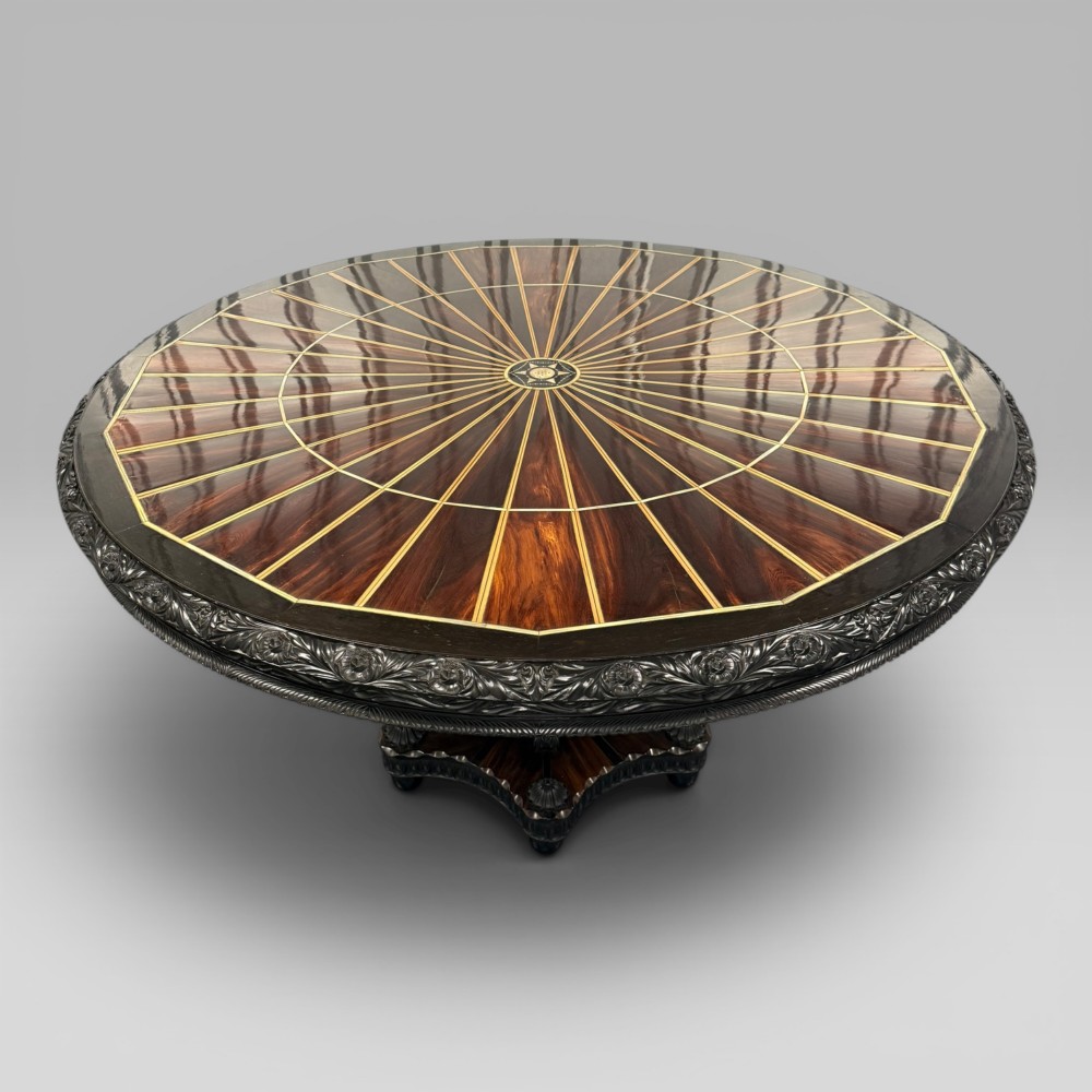 Overhead view of a circular centre table top showing radiating calamander wood veneers, light stringing, a central star motif, and a carved ebony border. View II