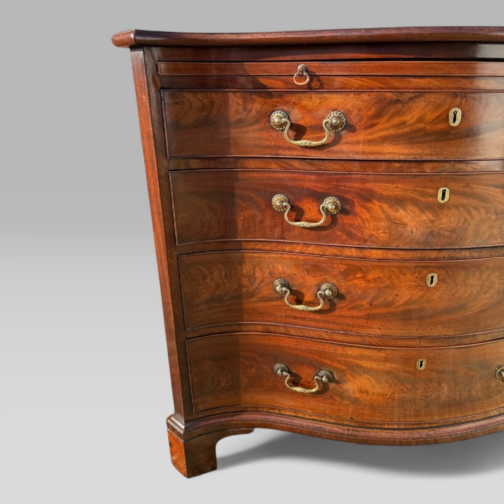 An 18th-century Georgian mahogany chest of drawers featuring a curved serpentine front, four graduated drawers, brass swan-neck handles, and a pull-out brushing slide. View IV