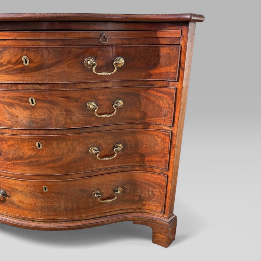 An 18th-century Georgian mahogany chest of drawers featuring a curved serpentine front, four graduated drawers, brass swan-neck handles, and a pull-out brushing slide. View V