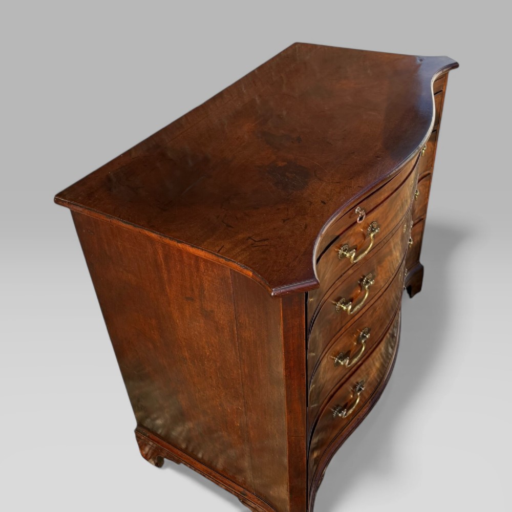 An 18th-century Georgian mahogany chest of drawers featuring a curved serpentine front, four graduated drawers, brass swan-neck handles, and a pull-out brushing slide. View VI