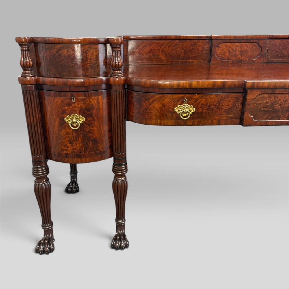 An antique Regency flame mahogany sideboard featuring a breakfront top, reeded columns, brass ring handles, and six carved lion paw feet. View VI