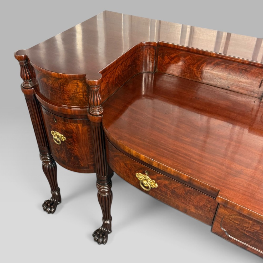 An antique Regency flame mahogany sideboard featuring a breakfront top, reeded columns, brass ring handles, and six carved lion paw feet. View VII