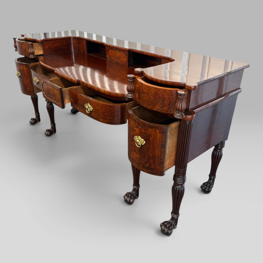An antique Regency flame mahogany sideboard featuring a breakfront top, reeded columns, brass ring handles, and six carved lion paw feet. View IX