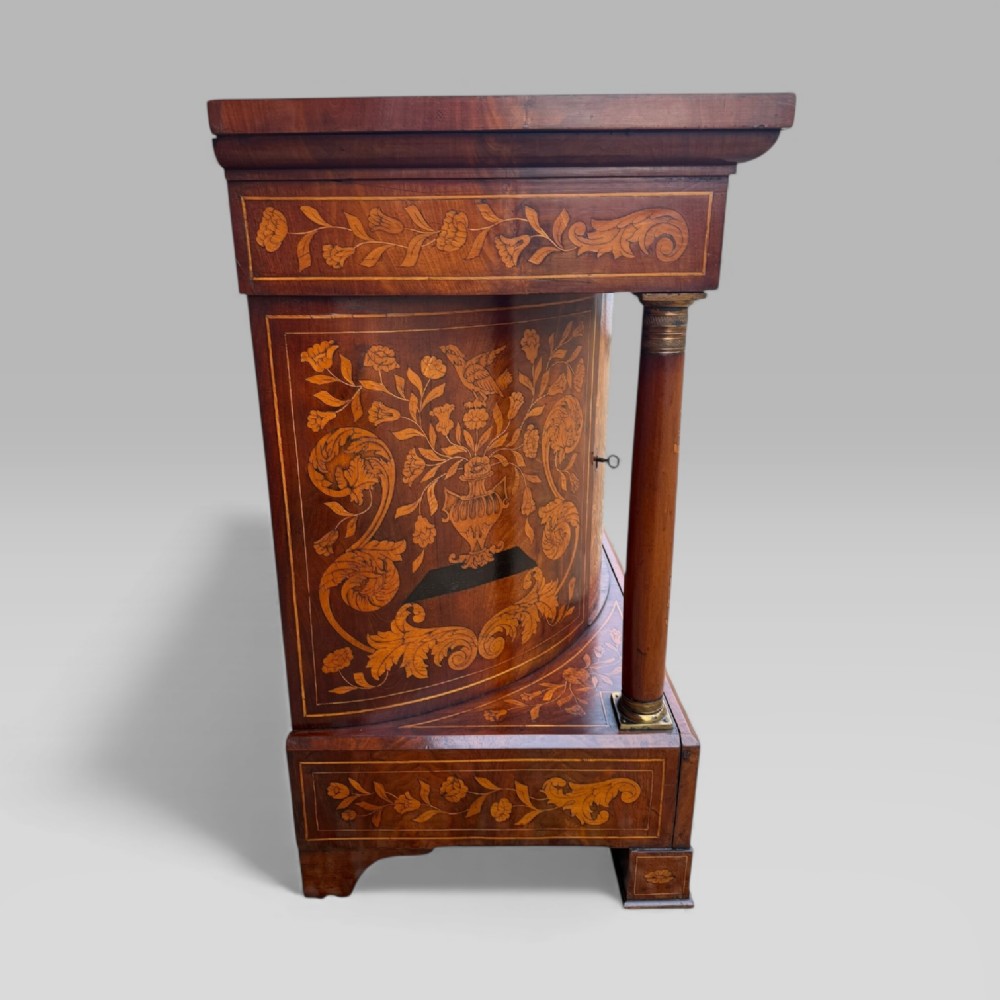 An antique Dutch mahogany console cabinet heavily decorated with intricate floral marquetry, featuring a single drawer above a rounded central cupboard door, flanked by turned columns. View VIII