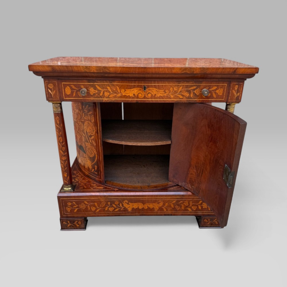 An antique Dutch mahogany console cabinet heavily decorated with intricate floral marquetry, featuring a single drawer above a rounded central cupboard door, flanked by turned columns. View XV