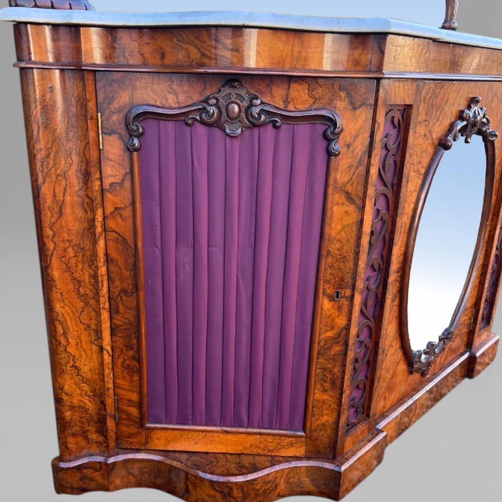 An antique Victorian burr walnut credenza featuring a white marble top, arched mirror back frame, central oval mirror door, and carved timber detailing. View VII