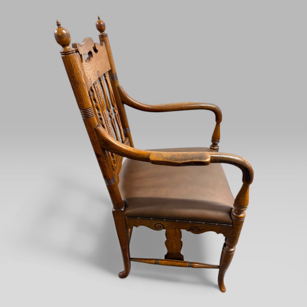 A Victorian golden oak armchair featuring a carved backrest, curved arms, brown leather seat with brass studs, and turned legs. View V