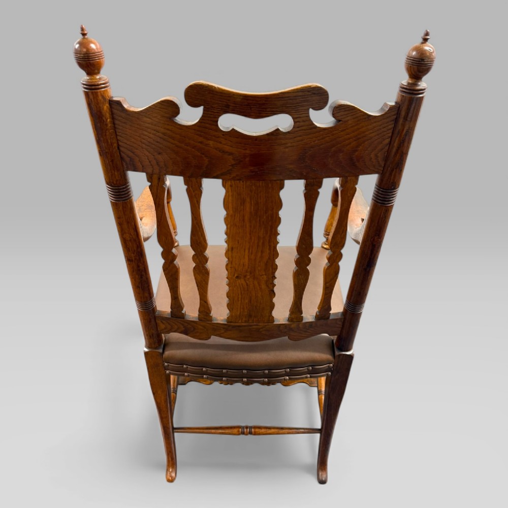 A Victorian golden oak armchair featuring a carved backrest, curved arms, brown leather seat with brass studs, and turned legs. View XI