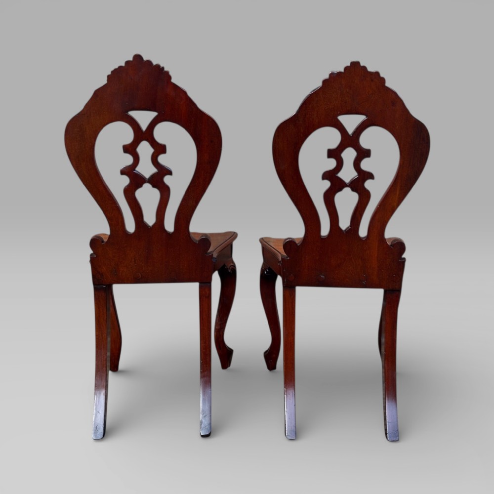 A pair of Victorian carved mahogany hall chairs featuring openwork pierced backs, shaped solid timber seats, and cabriole front legs. View VI
