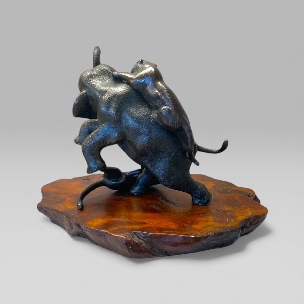 A patinated dark bronze sculpture depicting an elephant fighting off two tigers, mounted on a natural edge timber base. View IV