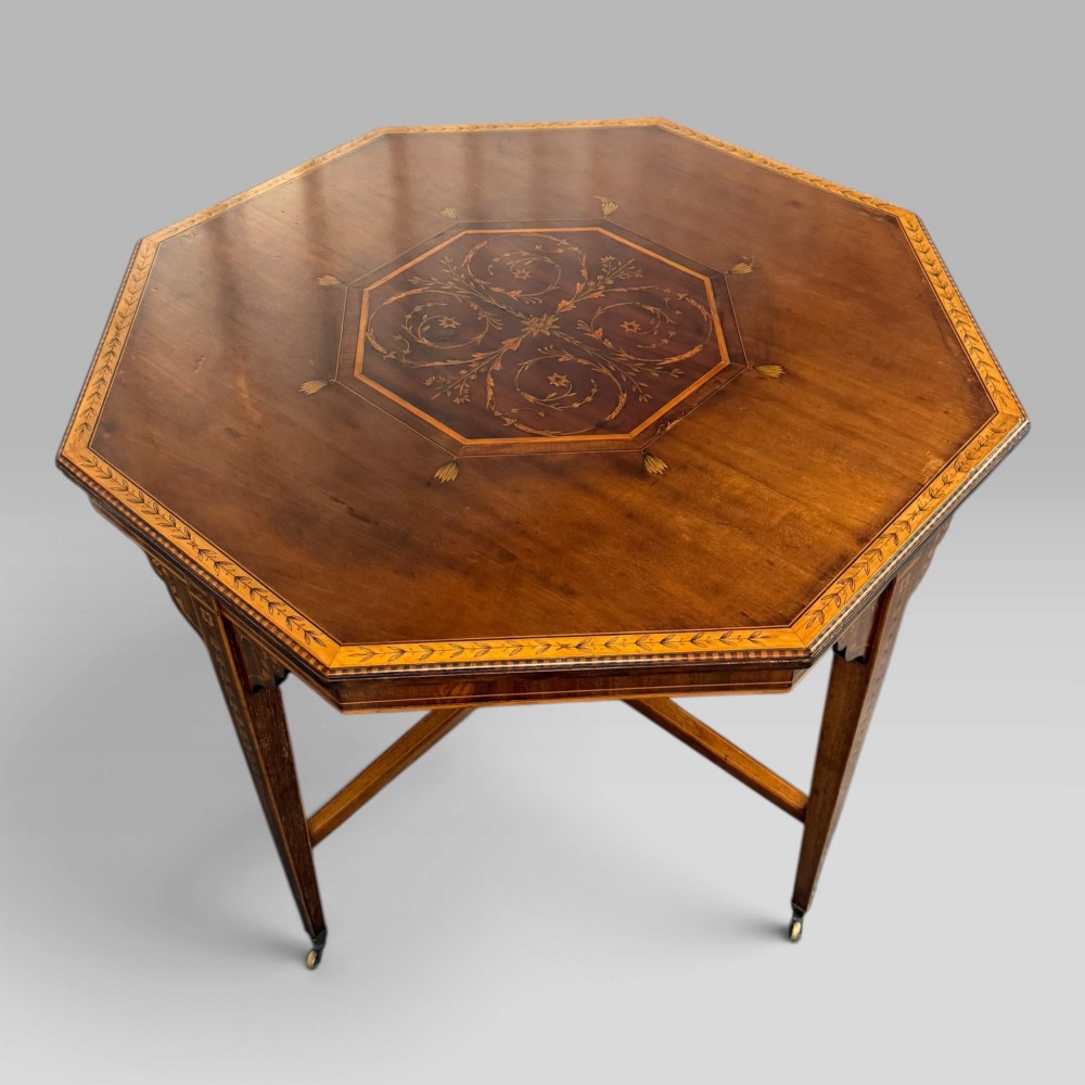 An octagonal Edwardian mahogany centre table featuring floral marquetry inlays, satinwood crossbanded borders, tapered legs, an X-stretcher, and brass castors.