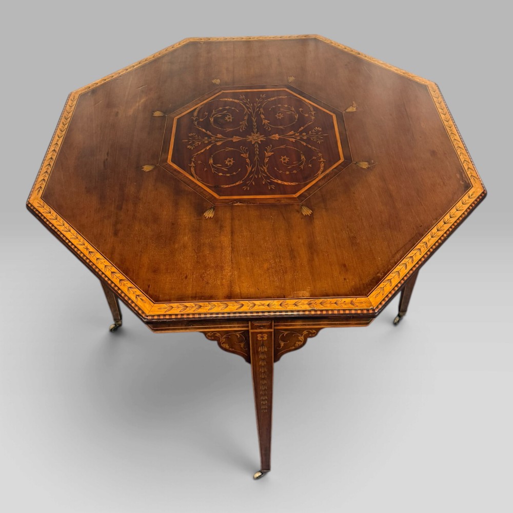 An octagonal Edwardian mahogany centre table featuring floral marquetry inlays, satinwood crossbanded borders, tapered legs, an X-stretcher, and brass castors. View II