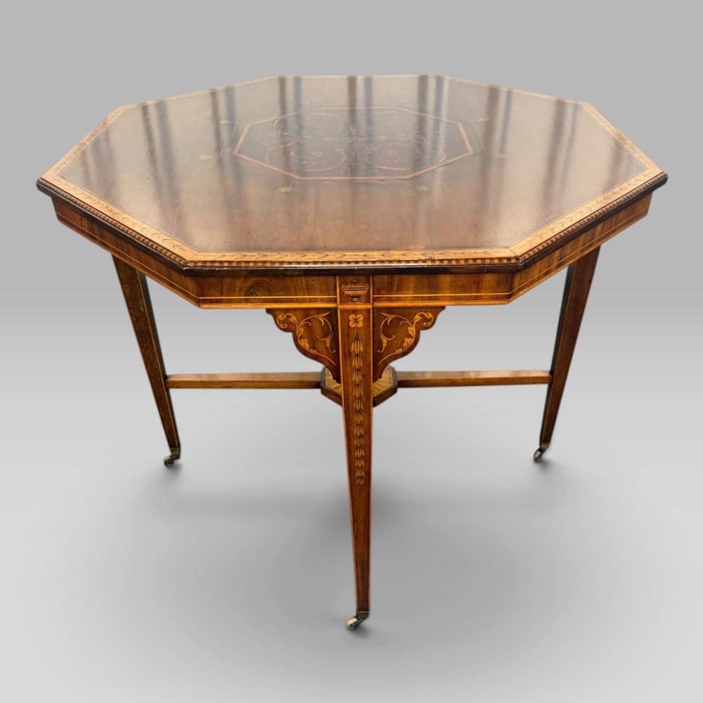 An octagonal Edwardian mahogany centre table featuring floral marquetry inlays, satinwood crossbanded borders, tapered legs, an X-stretcher, and brass castors. View IV