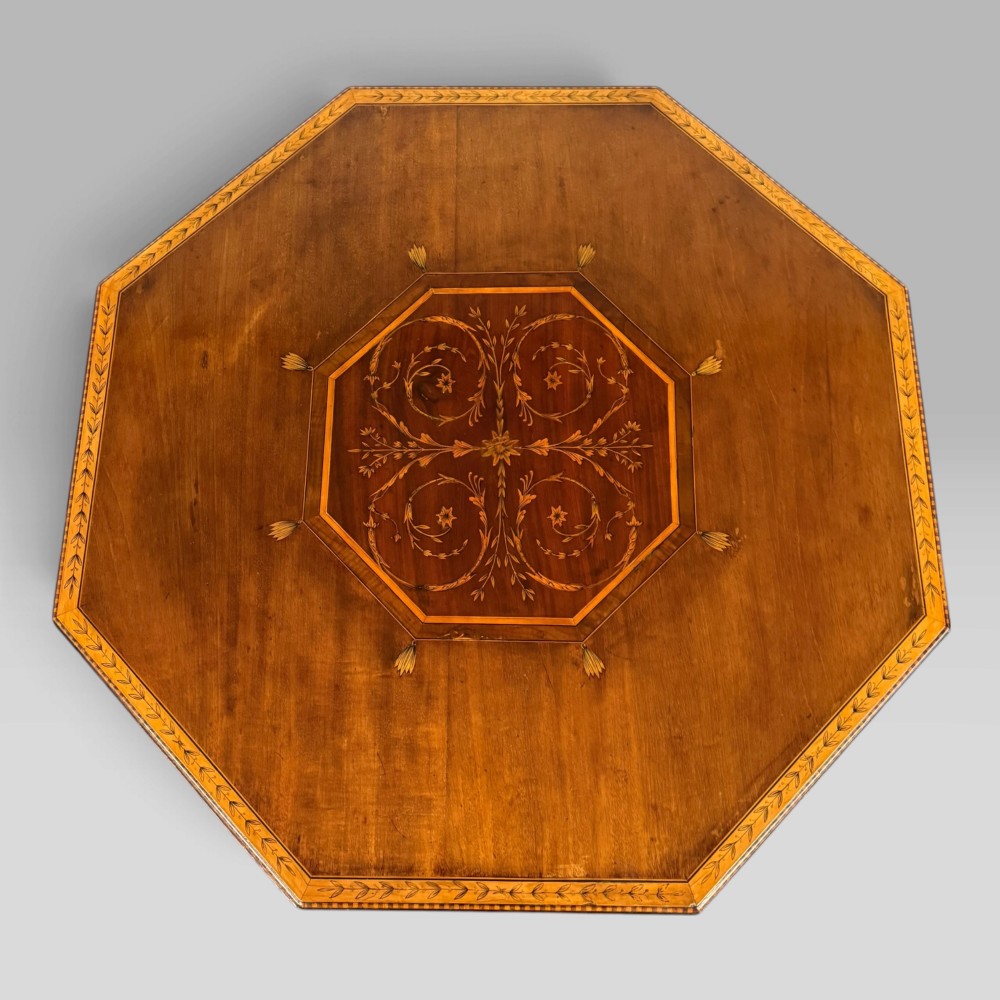 An octagonal Edwardian mahogany centre table featuring floral marquetry inlays, satinwood crossbanded borders, tapered legs, an X-stretcher, and brass castors. View V