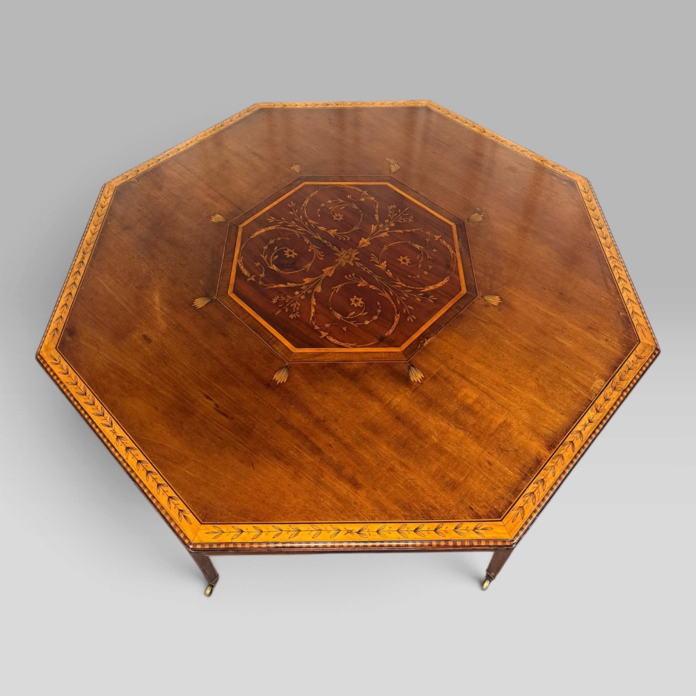An octagonal Edwardian mahogany centre table featuring floral marquetry inlays, satinwood crossbanded borders, tapered legs, an X-stretcher, and brass castors. View VI