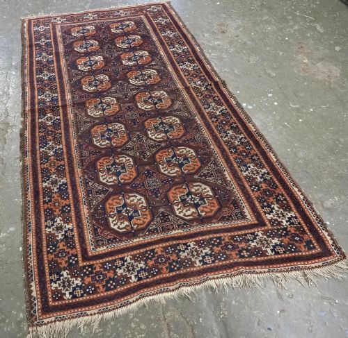 fig 1. An antique hand-knotted Baluch wool rug featuring repeating geometric octagonal medallions in terracotta, navy, and ivory across a dark brown field with intricate multi-banded borders.
