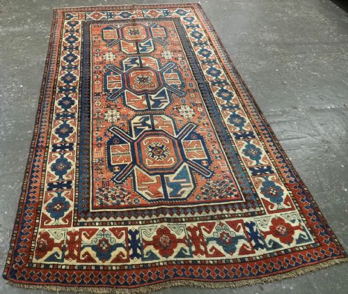fig 1. An antique hand-knotted Caucasian wool rug featuring three primary geometric medallions on a terracotta field with navy and cream patterned borders.