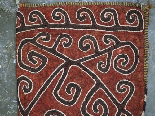 fig 1. A terracotta red felt panel embroidered with off-white and dark brown scroll motifs, featuring cascading tier tassels made of dark horsehair.