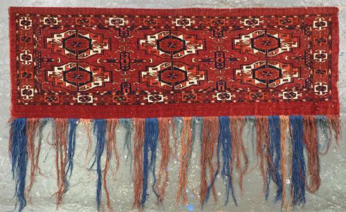 fig 1. A rectangular hand-woven Tekke Turkmen wool textile featuring six octagonal guls on a deep red field with an intricate geometric border and long blue and red fringe along the bottom edge.