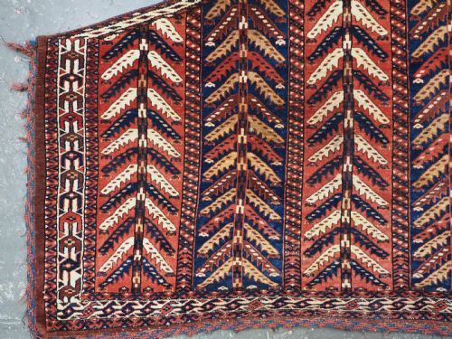 fig 2. A pentagonal antique Turkmen wool textile featuring vertical rows of white and gold chevron leaf motifs on a dark red background framed by geometric borders.