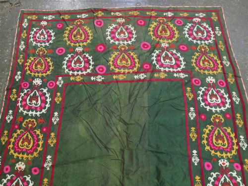 fig 1. An antique green silk suzani bed cover featuring a plain central panel surrounded by a broad embroidered border with magenta, yellow, and white floral motifs.