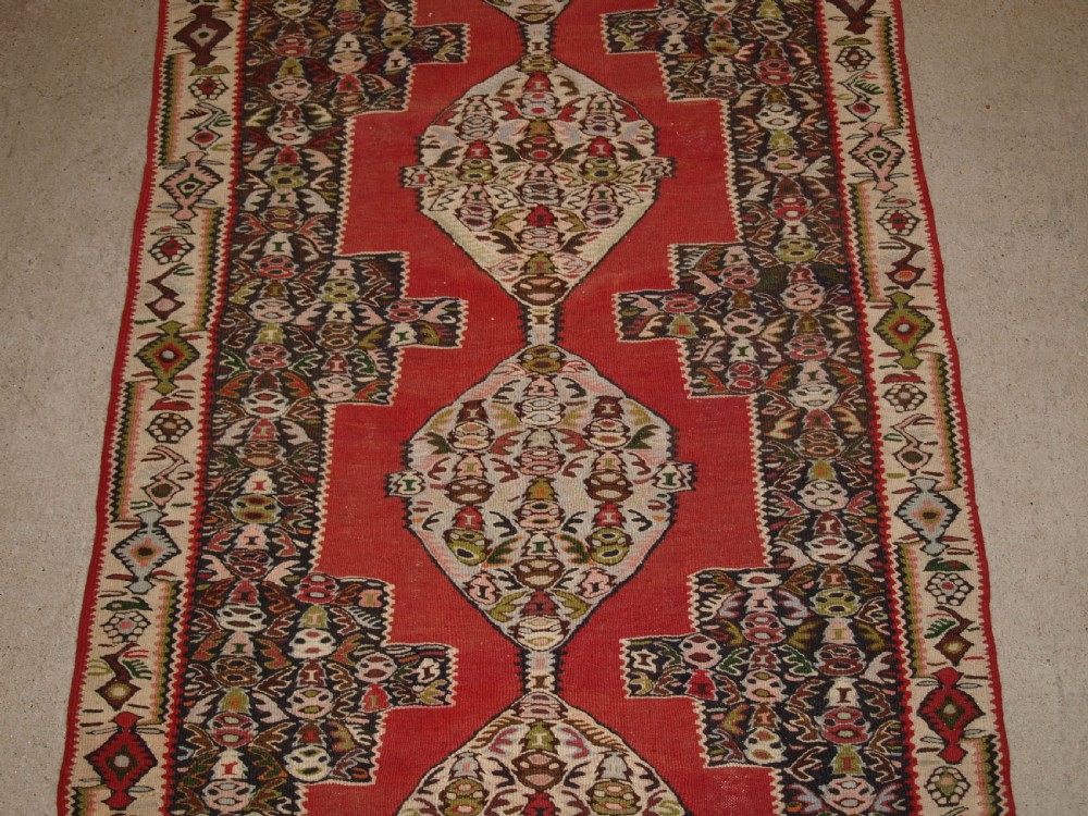 An antique Persian Senneh kilim runner featuring a sequence of linked diamond medallions on a vibrant red field with intricate geometric borders. View II