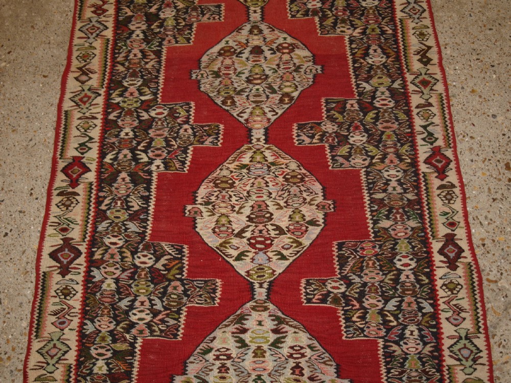 An antique Persian Senneh kilim runner featuring a sequence of linked diamond medallions on a vibrant red field with intricate geometric borders. View V