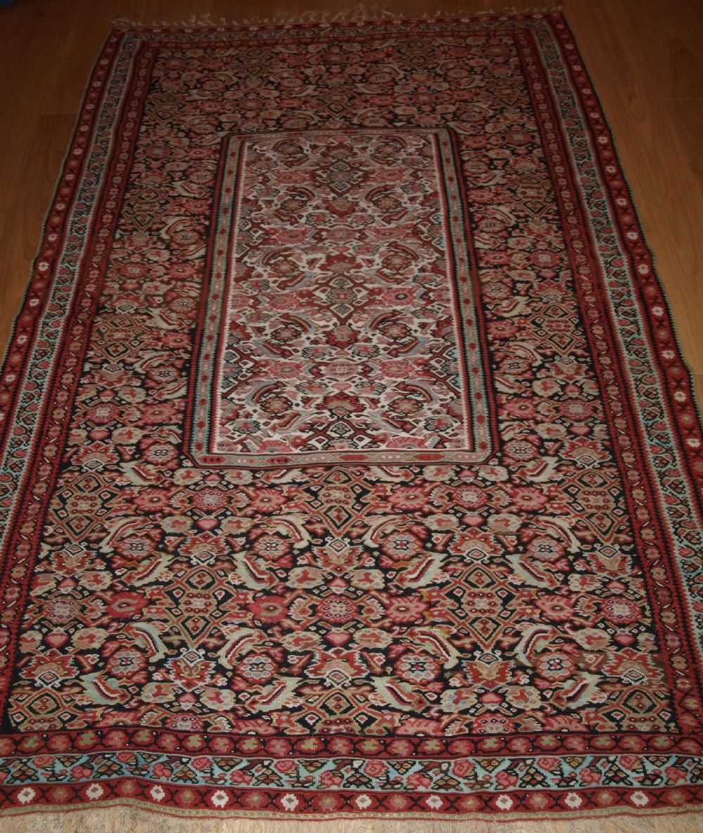 An antique flatwoven Persian Senneh kilim rug featuring a detailed central rectangle and surrounding border filled with intricate geometric and floral Herati patterns in red, ivory, and dark tones.