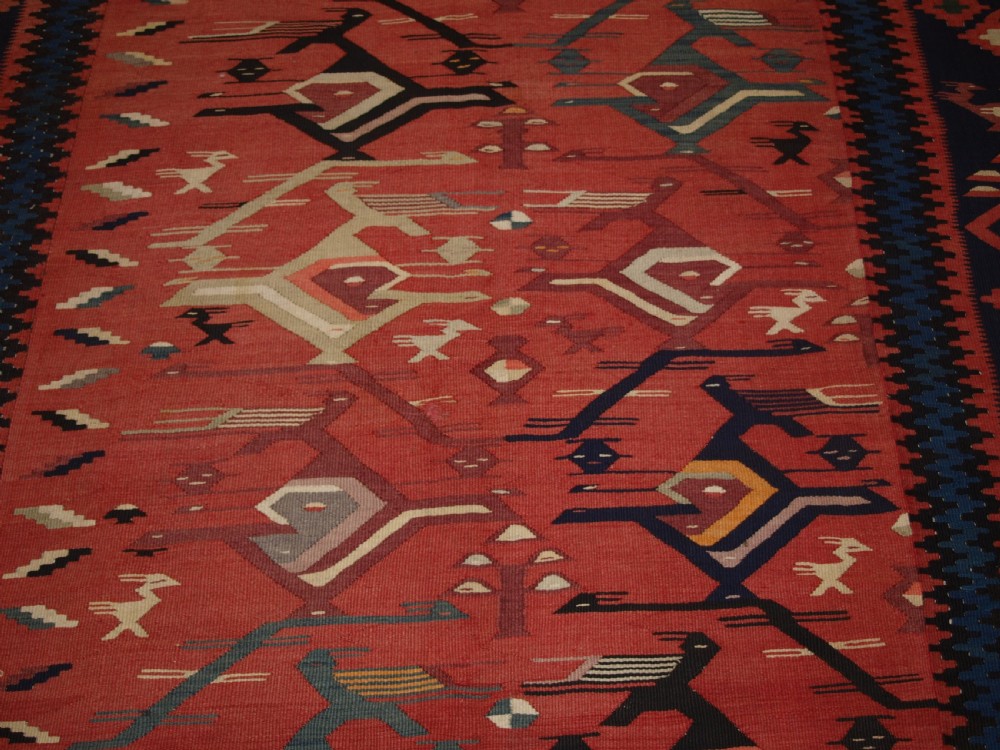 A large flatwoven Balkan Sharkoy kilim rug featuring terracotta red and navy blue borders decorated with stylised bird motifs and geometric patterns. View XII