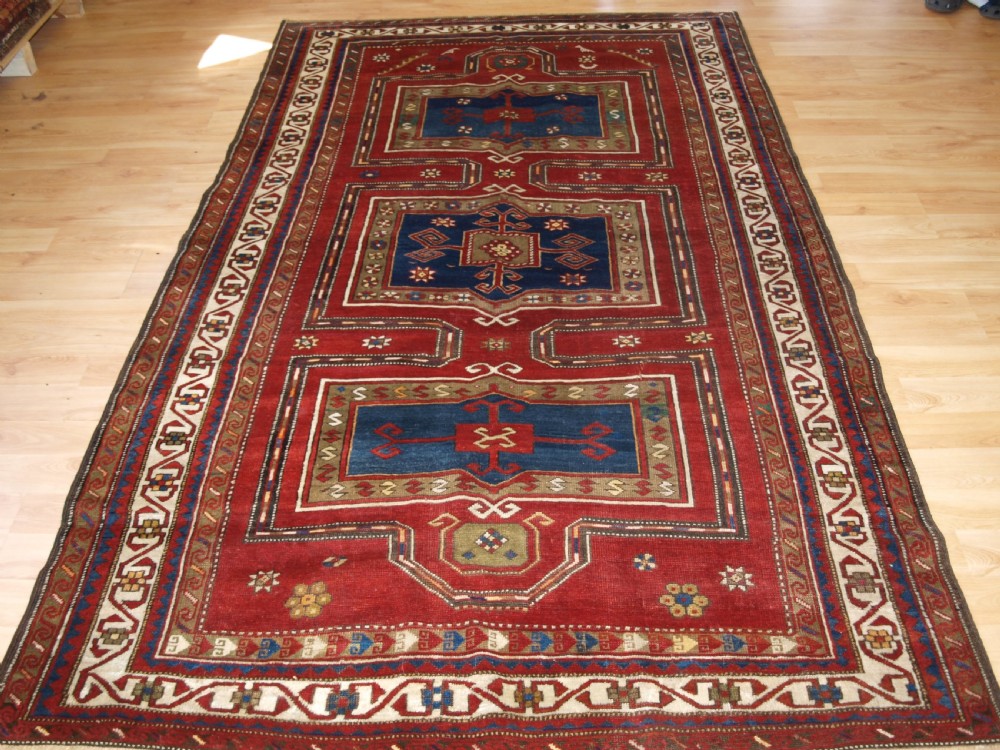 An antique Caucasian Fachralo Kazak hand-knotted wool long rug featuring a rich red field with three navy blue geometric medallions and elaborate borders.