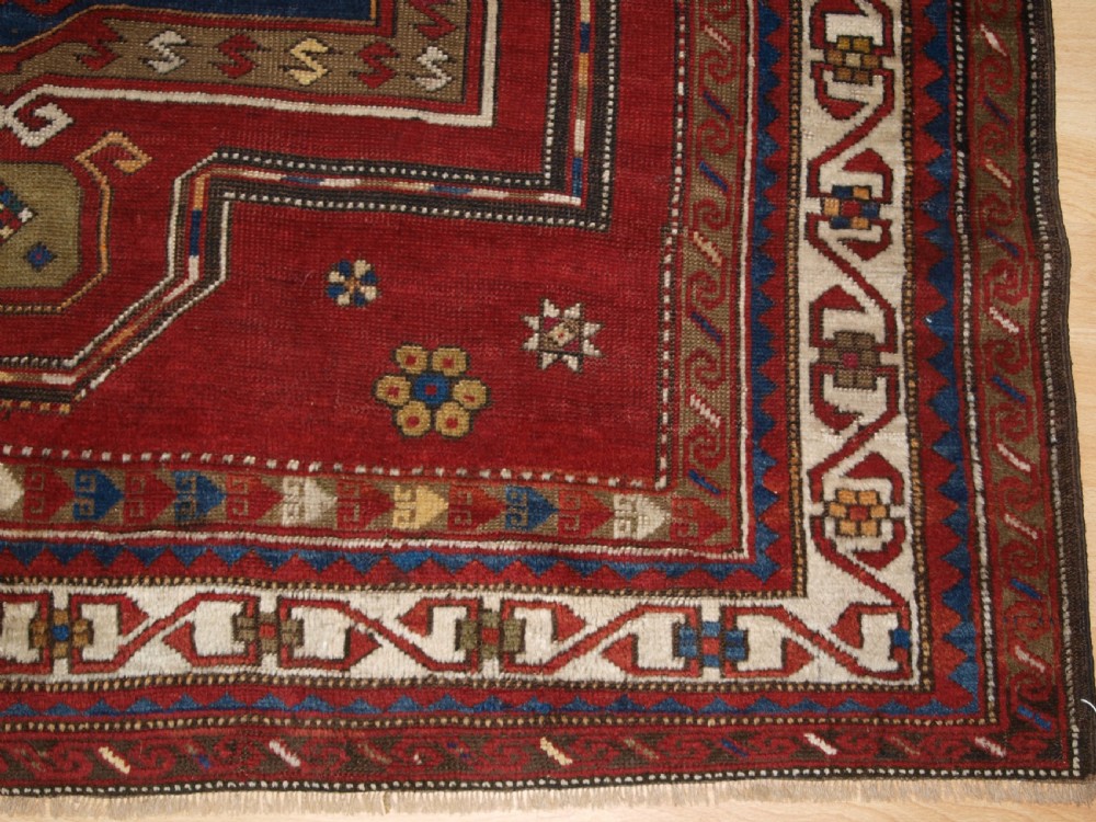 An antique Caucasian Fachralo Kazak hand-knotted wool long rug featuring a rich red field with three navy blue geometric medallions and elaborate borders. View X