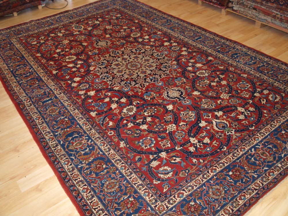 A large hand-knotted Persian Isfahan area rug featuring a red field filled with intricate floral arabesque vines and a central medallion surrounded by a dark blue ornate border.