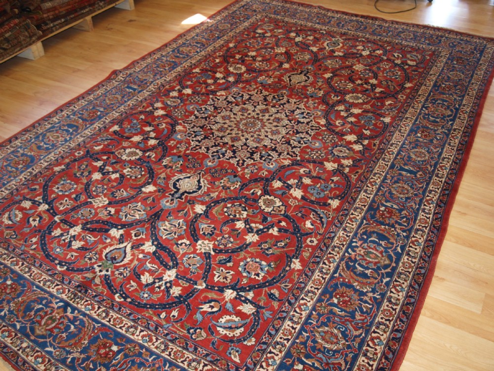 A large hand-knotted Persian Isfahan area rug featuring a red field filled with intricate floral arabesque vines and a central medallion surrounded by a dark blue ornate border. View II