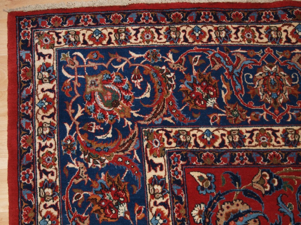 A large hand-knotted Persian Isfahan area rug featuring a red field filled with intricate floral arabesque vines and a central medallion surrounded by a dark blue ornate border. View III