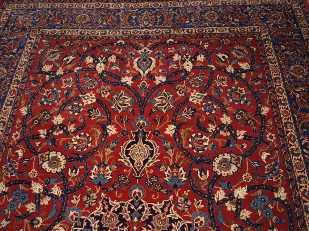 A large hand-knotted Persian Isfahan area rug featuring a red field filled with intricate floral arabesque vines and a central medallion surrounded by a dark blue ornate border. View IV