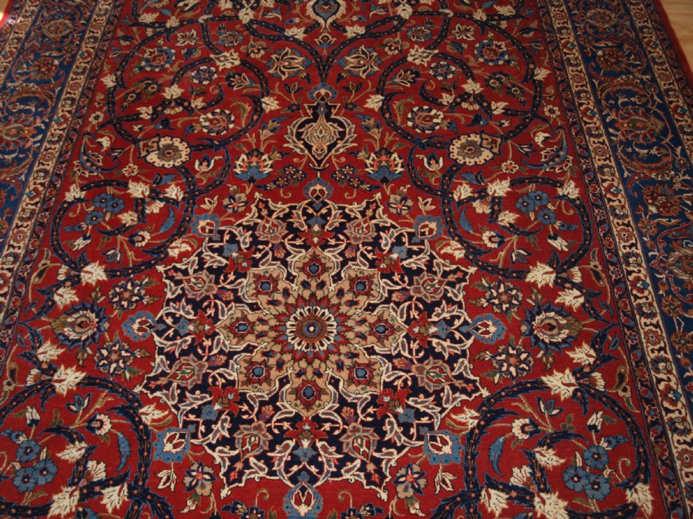 A large hand-knotted Persian Isfahan area rug featuring a red field filled with intricate floral arabesque vines and a central medallion surrounded by a dark blue ornate border. View V