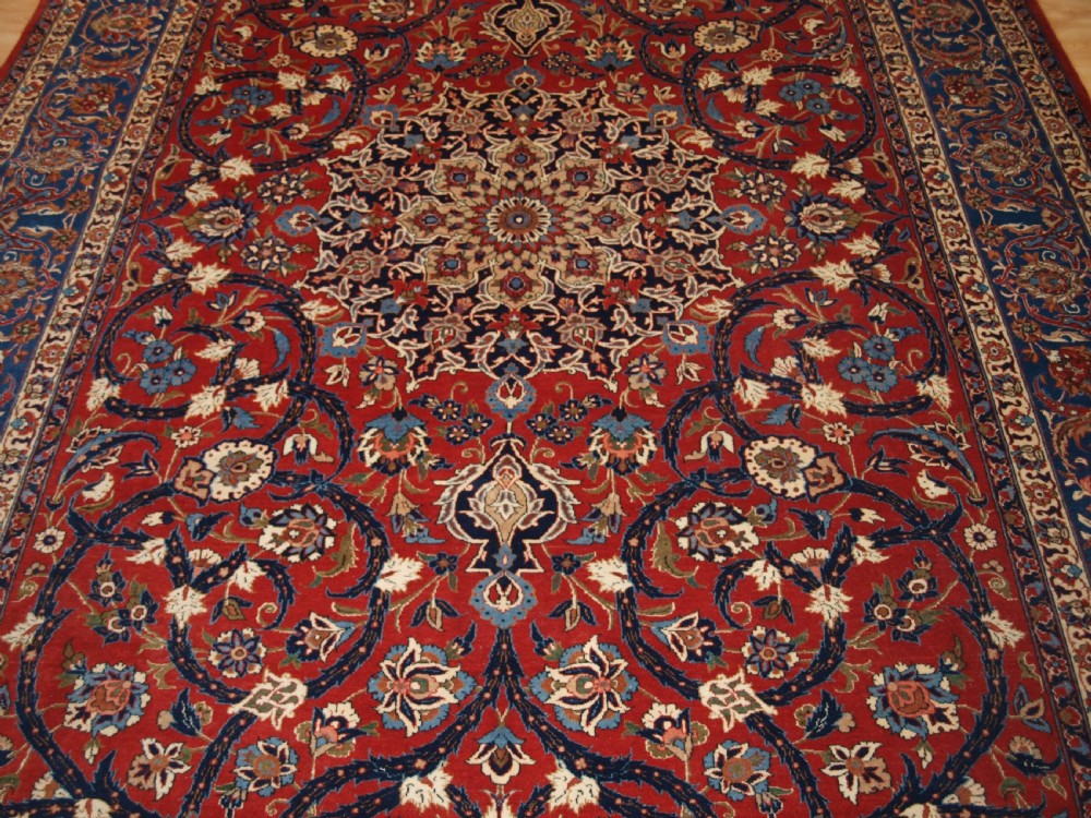 A large hand-knotted Persian Isfahan area rug featuring a red field filled with intricate floral arabesque vines and a central medallion surrounded by a dark blue ornate border. View VI