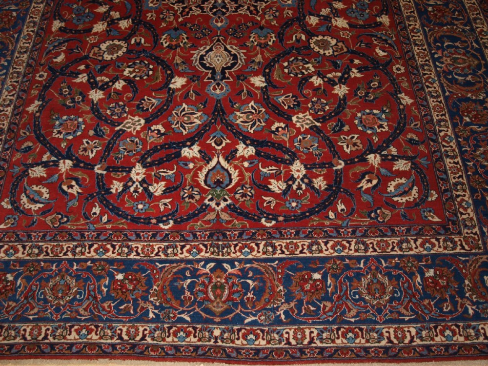 A large hand-knotted Persian Isfahan area rug featuring a red field filled with intricate floral arabesque vines and a central medallion surrounded by a dark blue ornate border. View VII