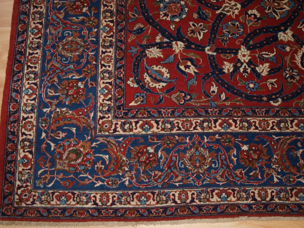 A large hand-knotted Persian Isfahan area rug featuring a red field filled with intricate floral arabesque vines and a central medallion surrounded by a dark blue ornate border. View VIII