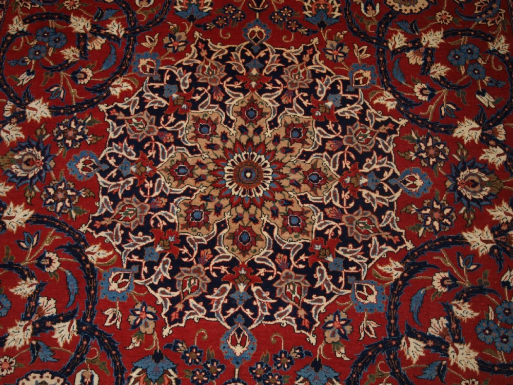 A large hand-knotted Persian Isfahan area rug featuring a red field filled with intricate floral arabesque vines and a central medallion surrounded by a dark blue ornate border. View IX